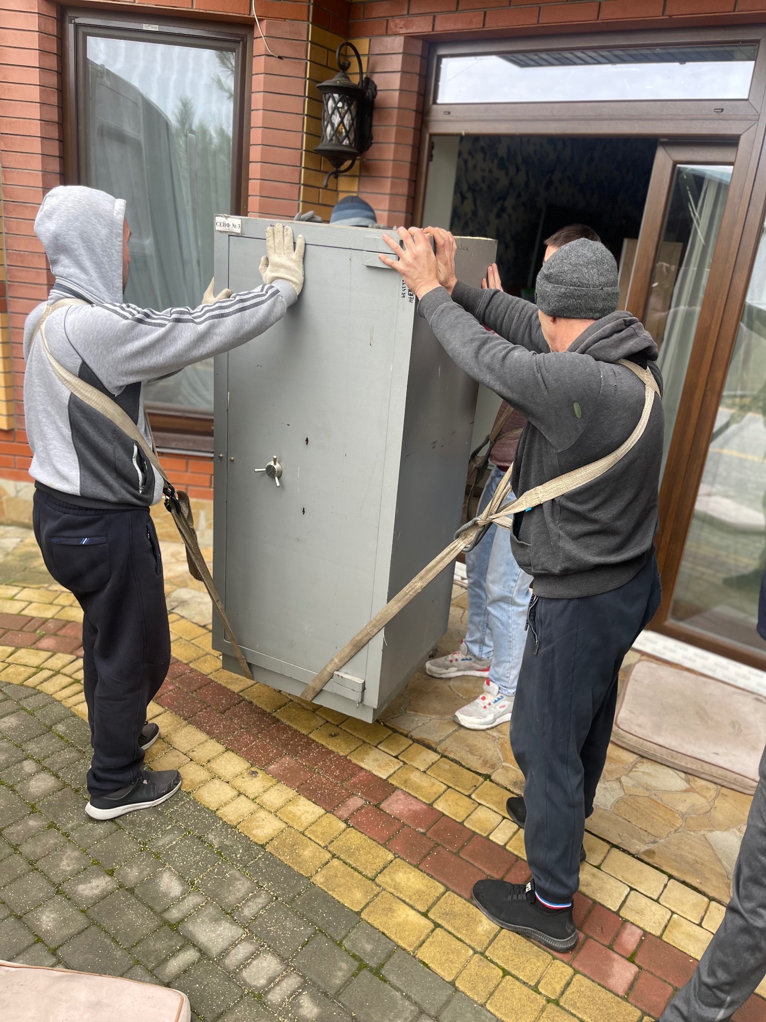 Trzech mężczyzn w roboczych ubraniach przenosi ciężki, szary sejf na zewnątrz budynku, używając pasów transportowych.