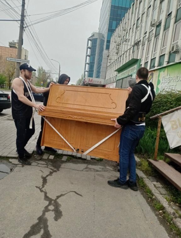 Trzech mężczyzn przenosi pianino po chodniku miejskim, zabezpieczone białymi pasami, na tle budynków biurowych i drzew.