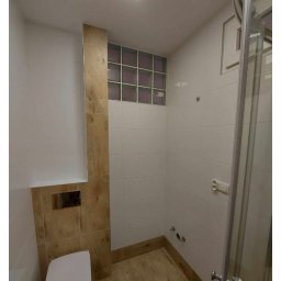 Kamil Gabriel - Nowoczesna łazienka po remoncie z kabiną prysznicową, podwieszaną toaletą, wykończeniem w bieli i drewnie oraz oknem z luksferów.