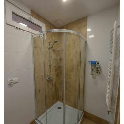 Kamil Gabriel - Wykończona łazienka w Bytomiu z kabiną prysznicową z zaokrąglonymi szybami, beżowymi płytkami imitującymi drewno, białymi płytkami na ścianach i drewnianą podłogą, widoczny filtr wody.