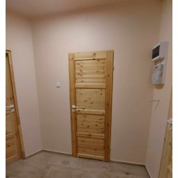 Kamil Gabriel - Wnętrze z dwiema drewnianymi, surowymi drzwiami z poziomymi panelami, jasnymi ścianami i podłogą z płytek imitujących drewno.