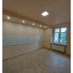 Pusty pokój po remoncie z panelami podłogowymi imitującymi drewno, pomalowanymi ścianami na jasny beż i listwą z białymi gniazdkami elektrycznymi na ścianie, oświetlony światłem z okna i lampami...