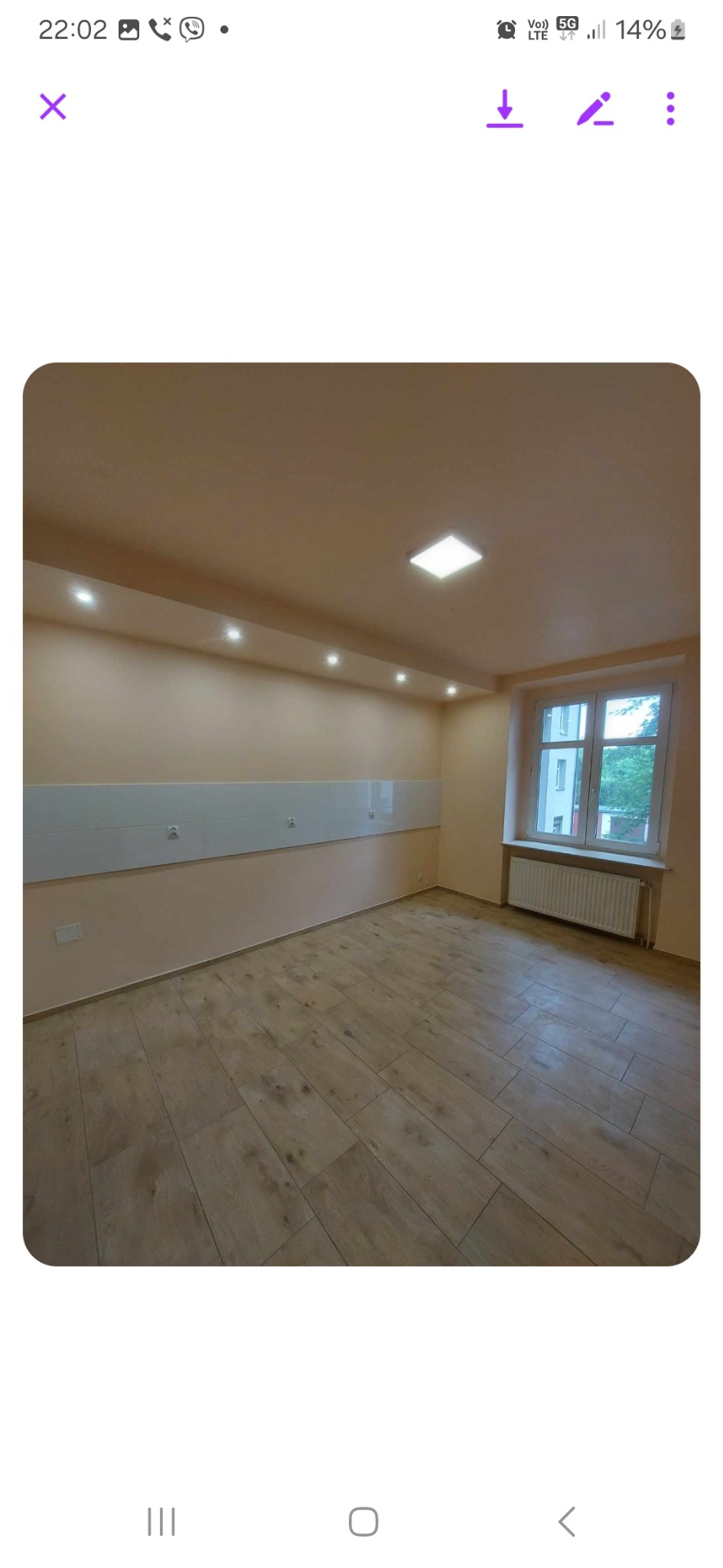 Pusty pokój po remoncie z panelami podłogowymi imitującymi drewno, białymi płytkami na ścianie, grzejnikiem pod oknem i oświetleniem sufitowym.