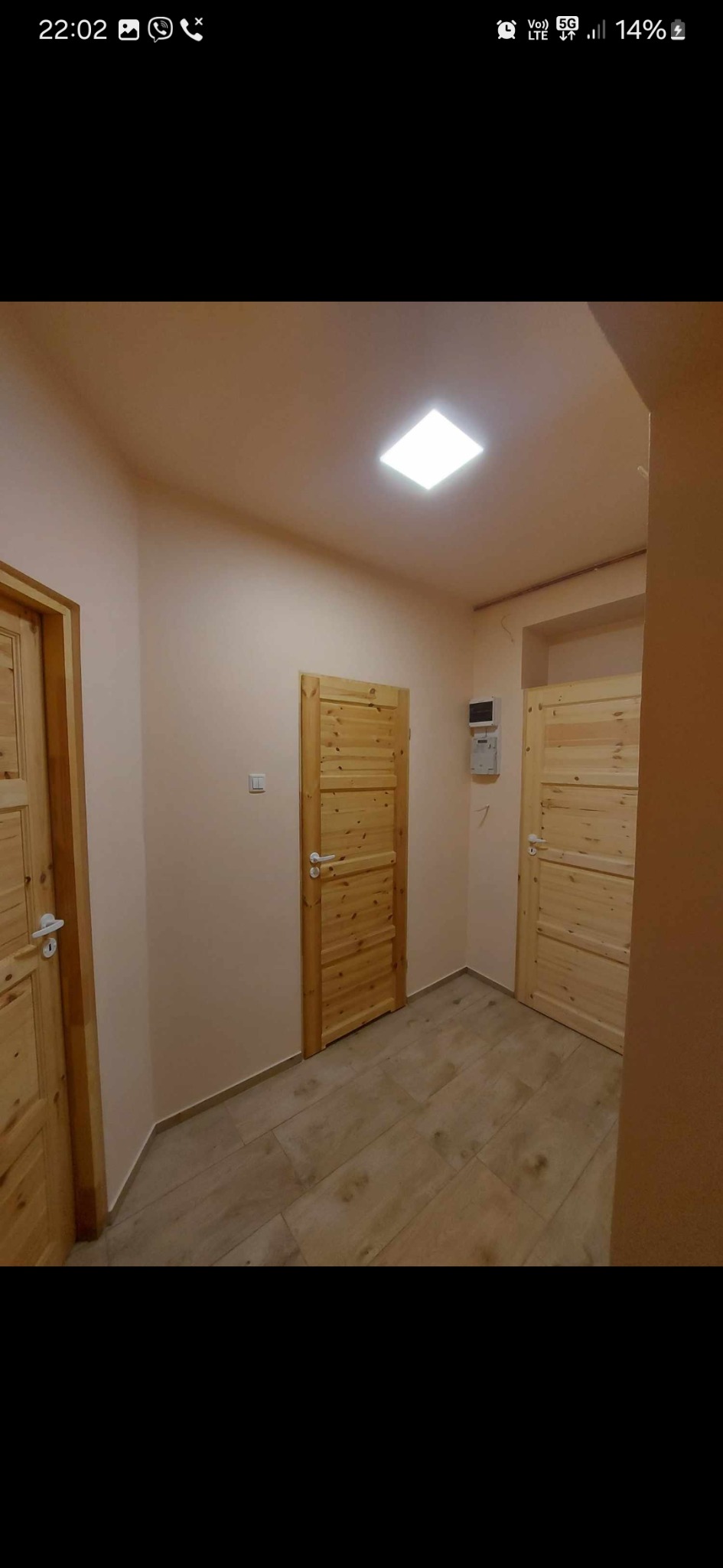 Wnętrze korytarza po remoncie z trzema drewnianymi drzwiami, beżowymi ścianami i podłogą imitującą drewno, oświetlone centralnym kwadratowym panelem LED.