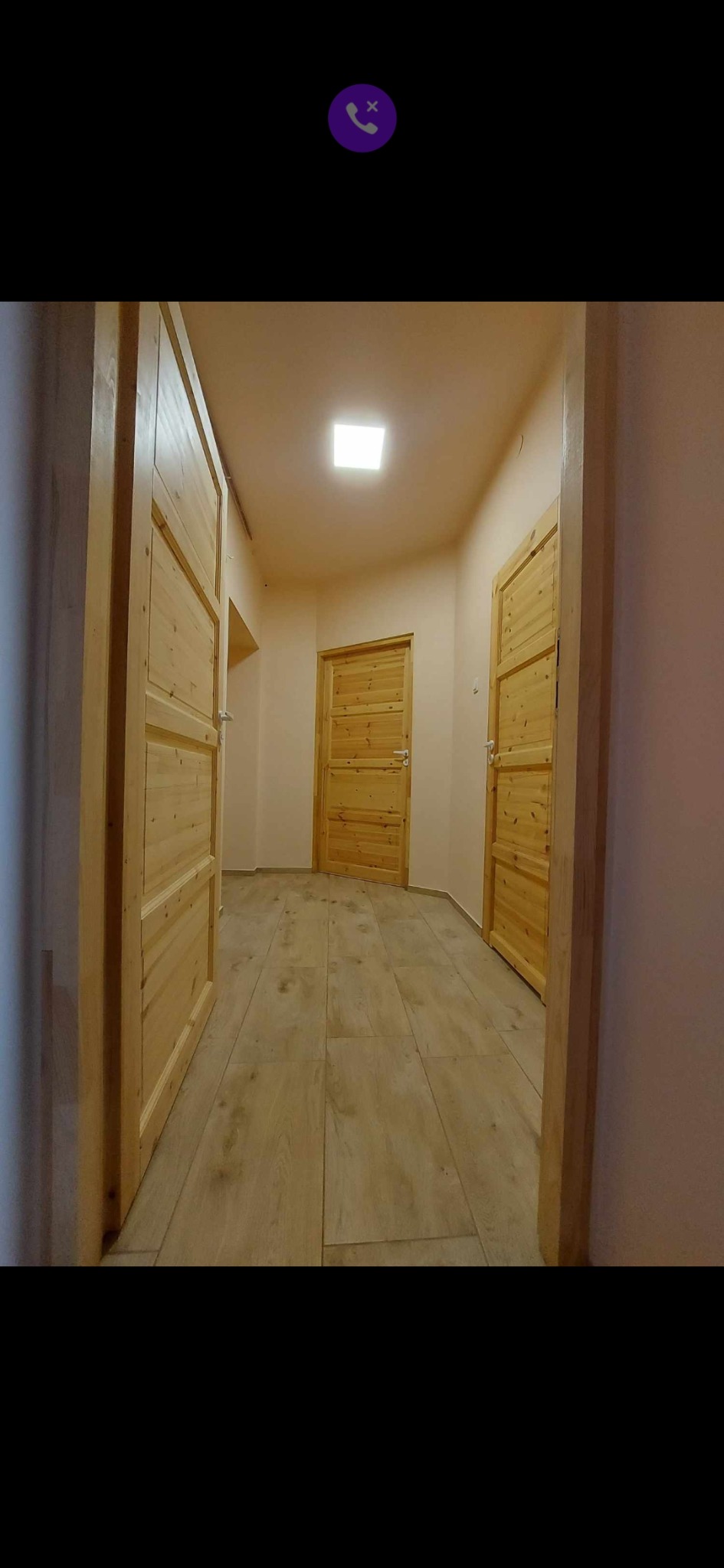 Korytarz z trzema drewnianymi drzwiami o panelowej konstrukcji i jasną podłogą imitującą drewno, oświetlony kwadratową lampą sufitową.