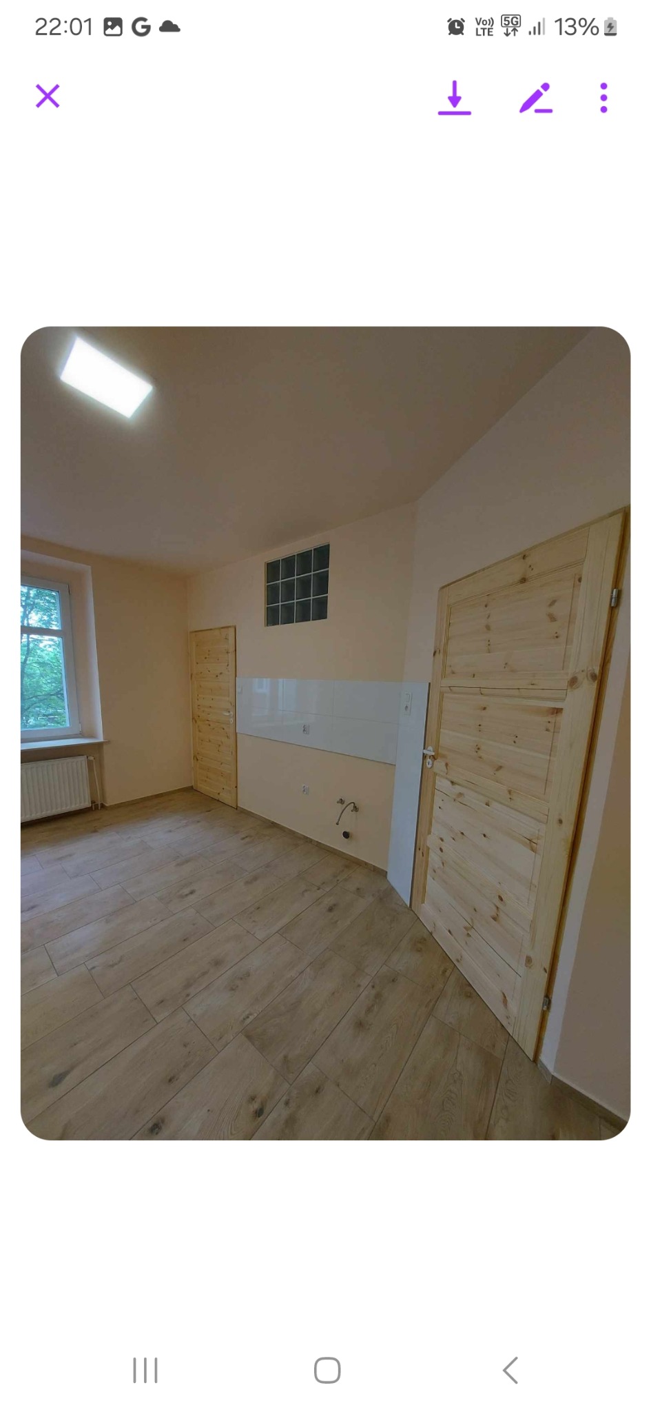 Jasne wnętrze po remoncie: drewnopodobna podłoga, dwie surowe drewniane drzwi z poziomymi panelami, białe płytki nad blatem w kuchni, okno z widokiem na zieleń.