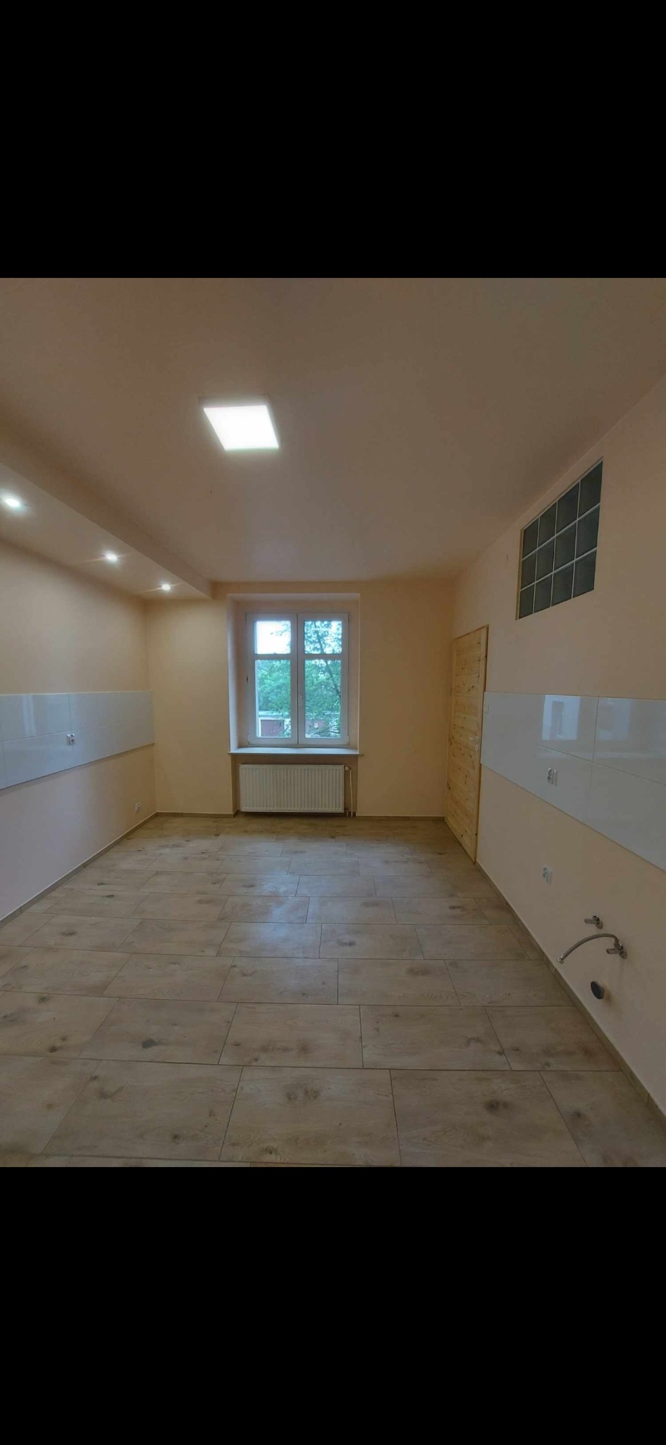 Puste pomieszczenie po remoncie z nową podłogą z paneli imitujących drewno, białymi płytkami na ścianach do połowy wysokości i oknem z widokiem na zieleń.