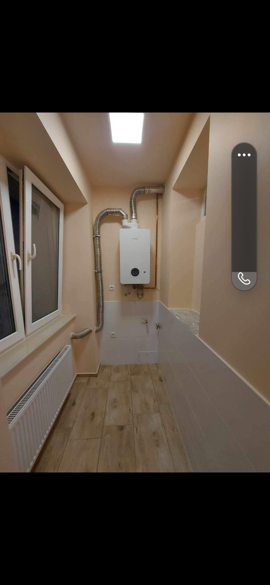 Biały piec gazowy kondensacyjny zamontowany na ścianie w nowo wyremontowanym pomieszczeniu z drewnopodobną podłogą, grzejnikiem pod oknem i częściowo wykafelkowanymi ścianami.