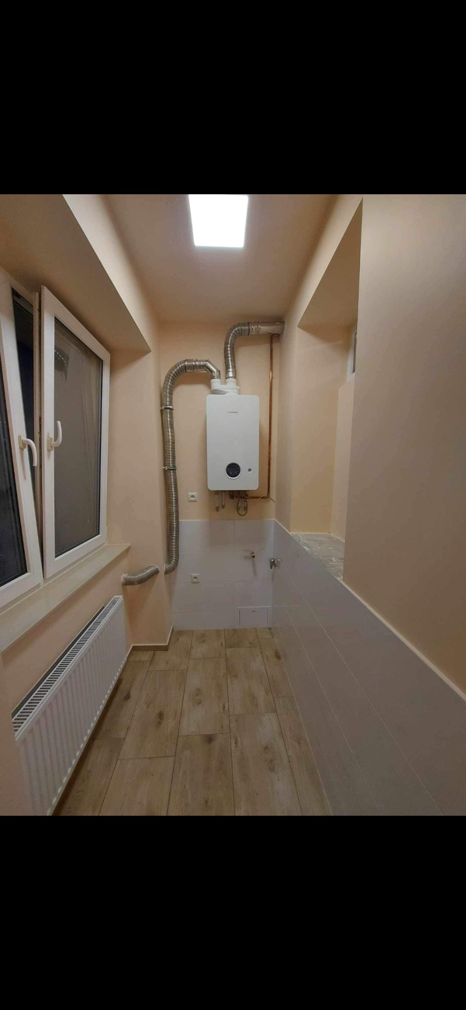 Wnętrze wyremontowanej łazienki z drewnopodobną podłogą, białymi płytkami na ścianach do połowy wysokości i zamontowanym piecem gazowym z odprowadzeniem spalin.