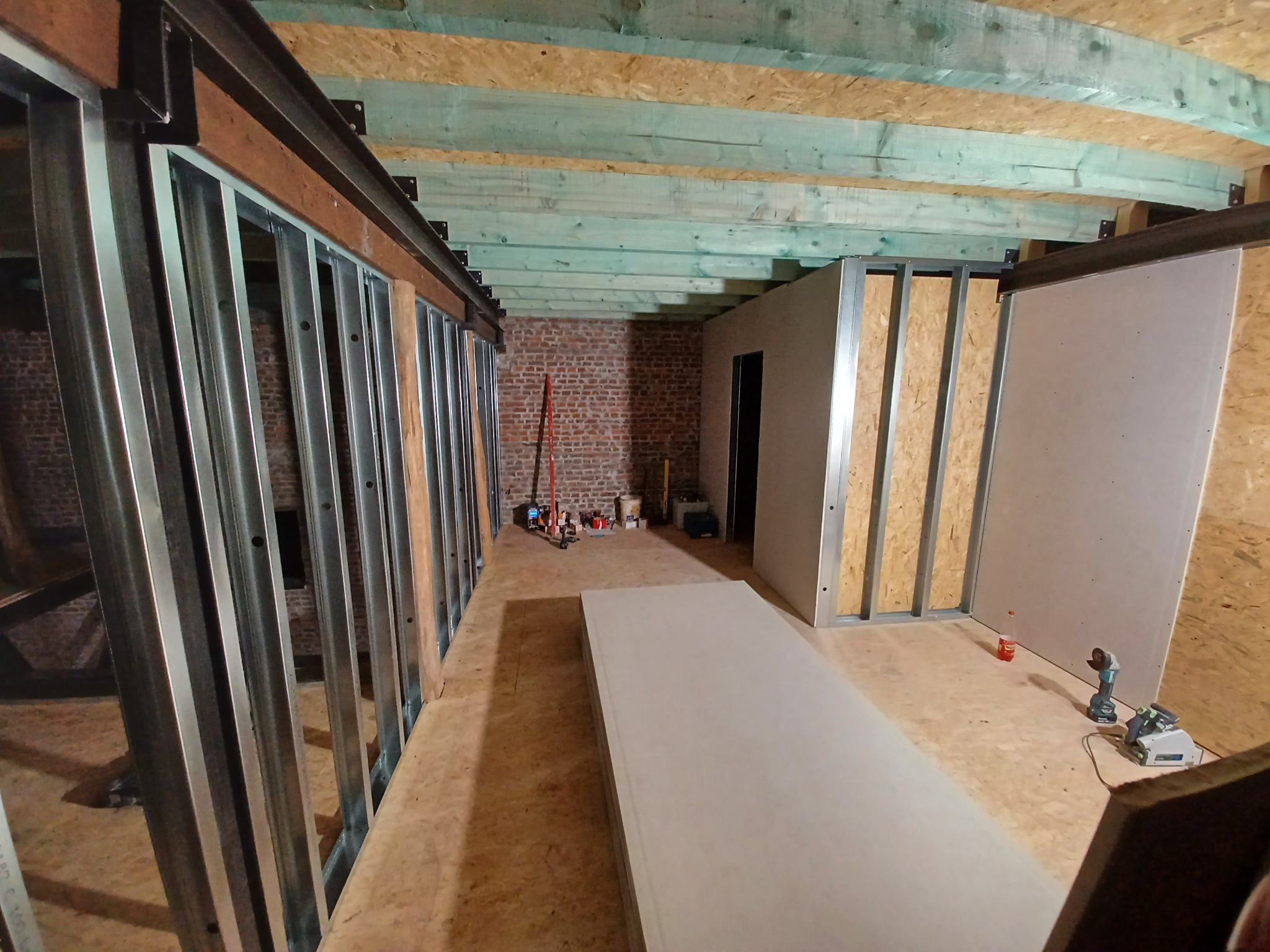 Wnętrze poddasza w trakcie adaptacji: metalowe profile ścian działowych, drewniana konstrukcja dachu z widocznymi belkami, przygotowane płyty karton-gips i narzędzia budowlane na podłodze.