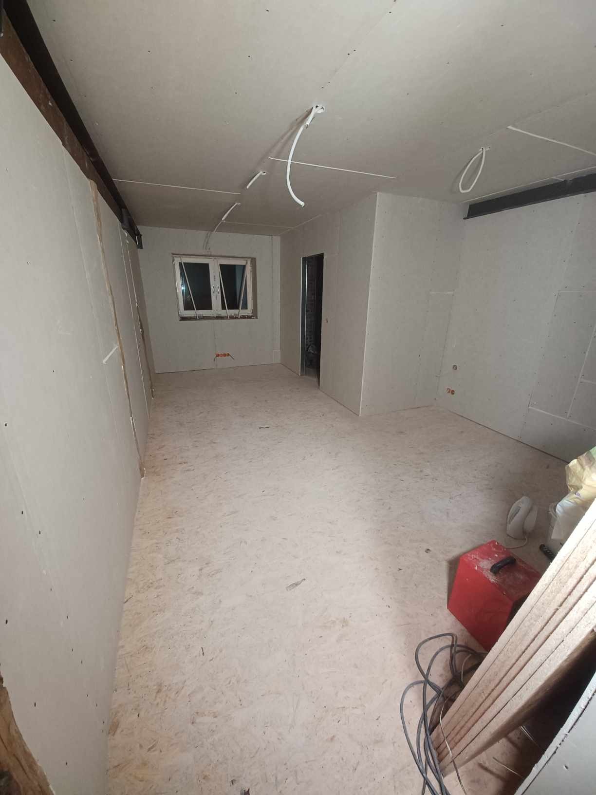 Wnętrze w trakcie prac wykończeniowych: surowe ściany z płyt gipsowo-kartonowych, drewniana podłoga z widocznym wzorem, wystające przewody elektryczne, okno z widokiem na zewnątrz oraz narzędzia...