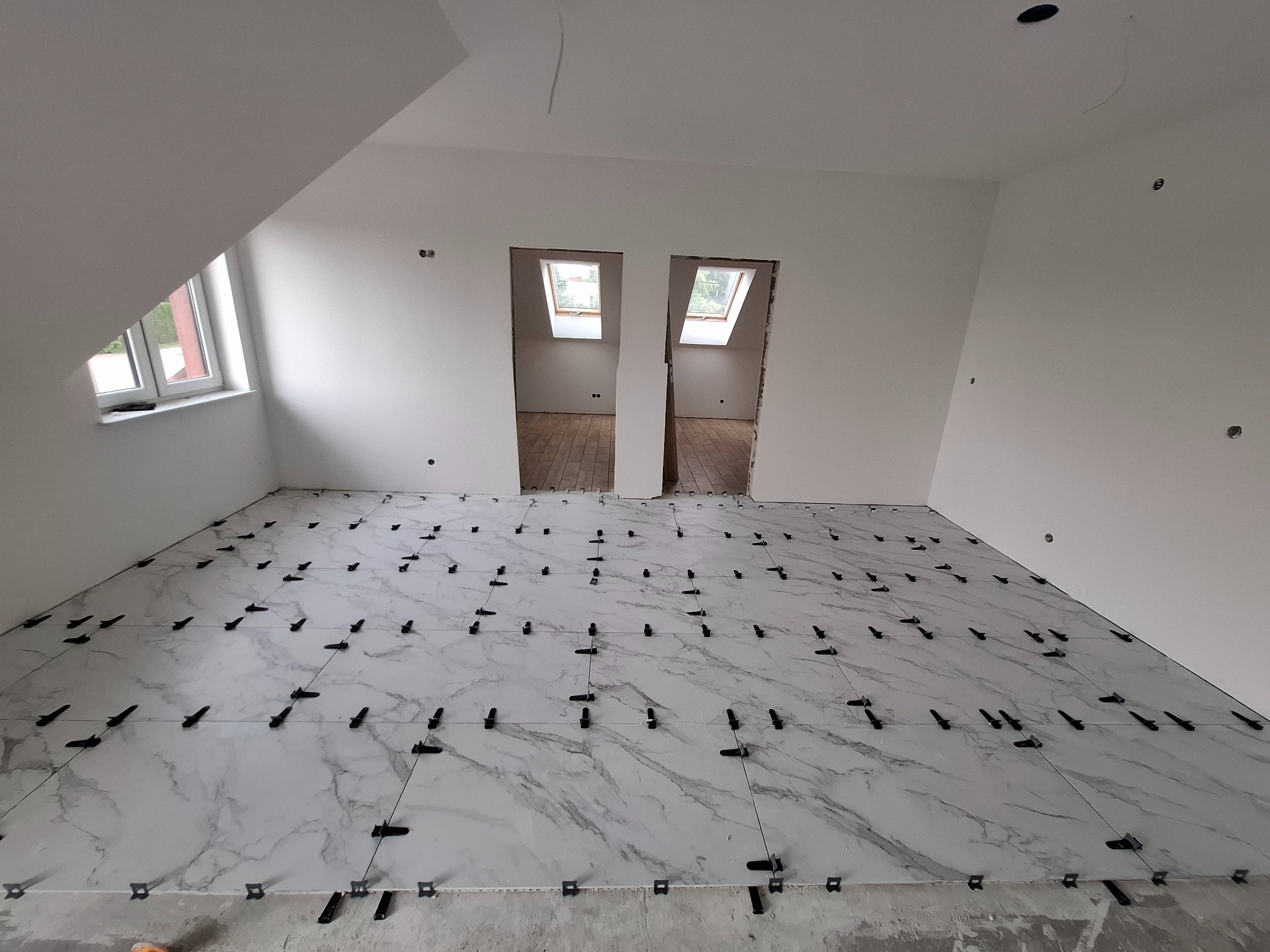 Poddasze z oknami dachowymi, białe ściany, podłoga w trakcie układania płytek imitujących marmur, z użyciem czarnych klipsów poziomujących.