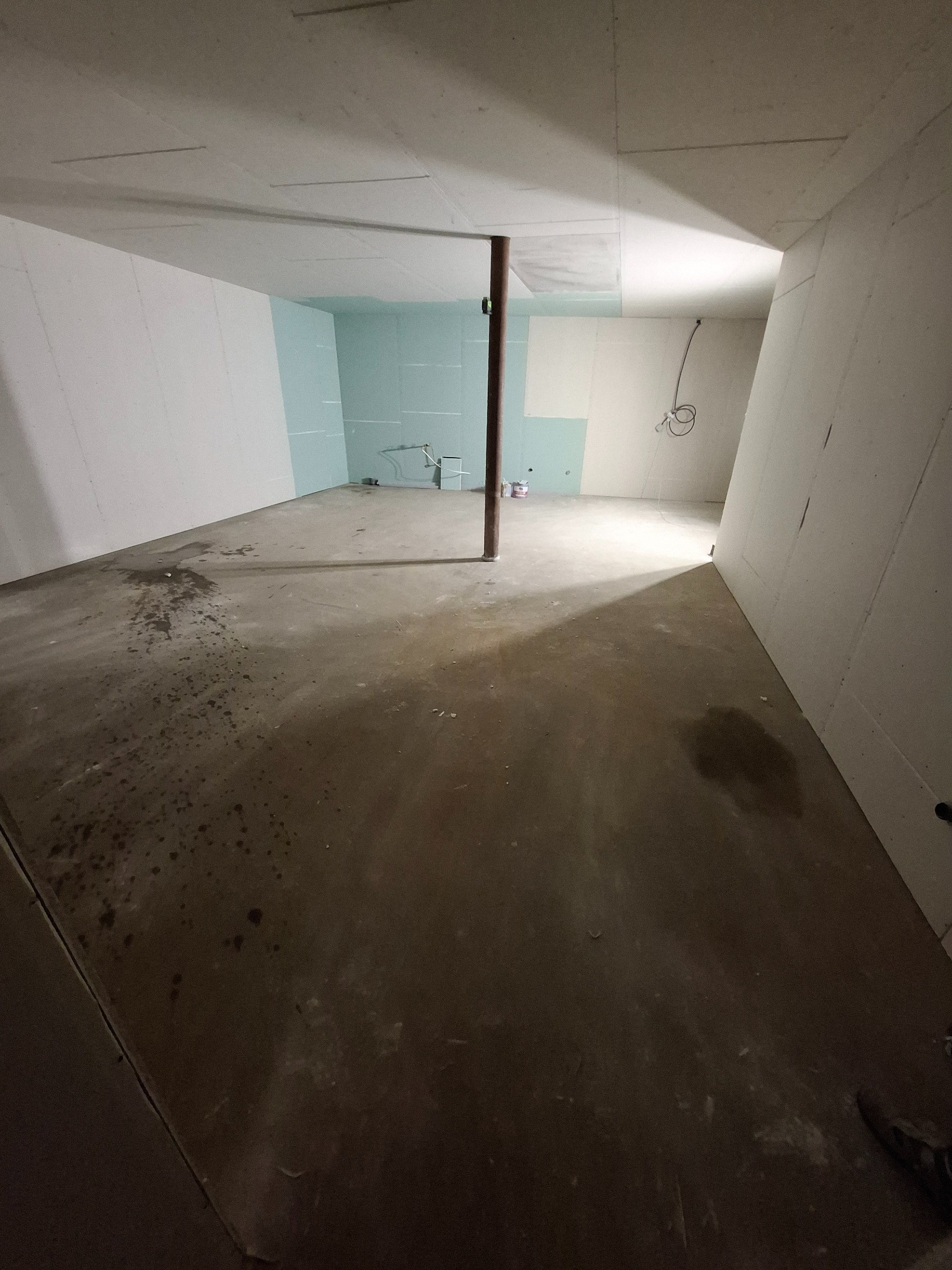 Surowe wnętrze w trakcie prac wykończeniowych: ściany częściowo pokryte płytami gipsowo-kartonowymi, surowy beton na podłodze z plamami, widoczna instalacja elektryczna, drewniany słup...