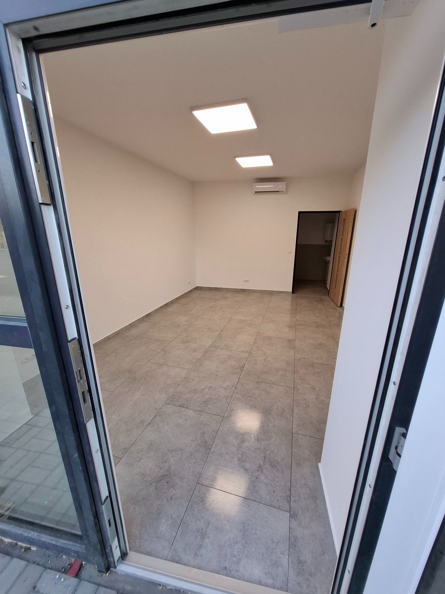 Pusty pokój z szarymi płytkami podłogowymi, widziany z perspektywy otwartych szklanych drzwi wejściowych; na ścianie klimatyzator i kwadratowe lampy sufitowe.