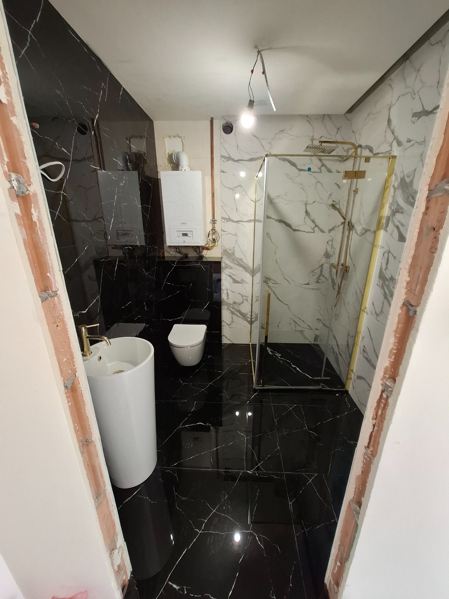 Łazienka w trakcie remontu: czarne marmurowe płytki na podłodze i ścianie, biała umywalka na postumencie z złotą armaturą, kabina prysznicowa z złotymi ramami, wiszący biały bojler, widoczne...