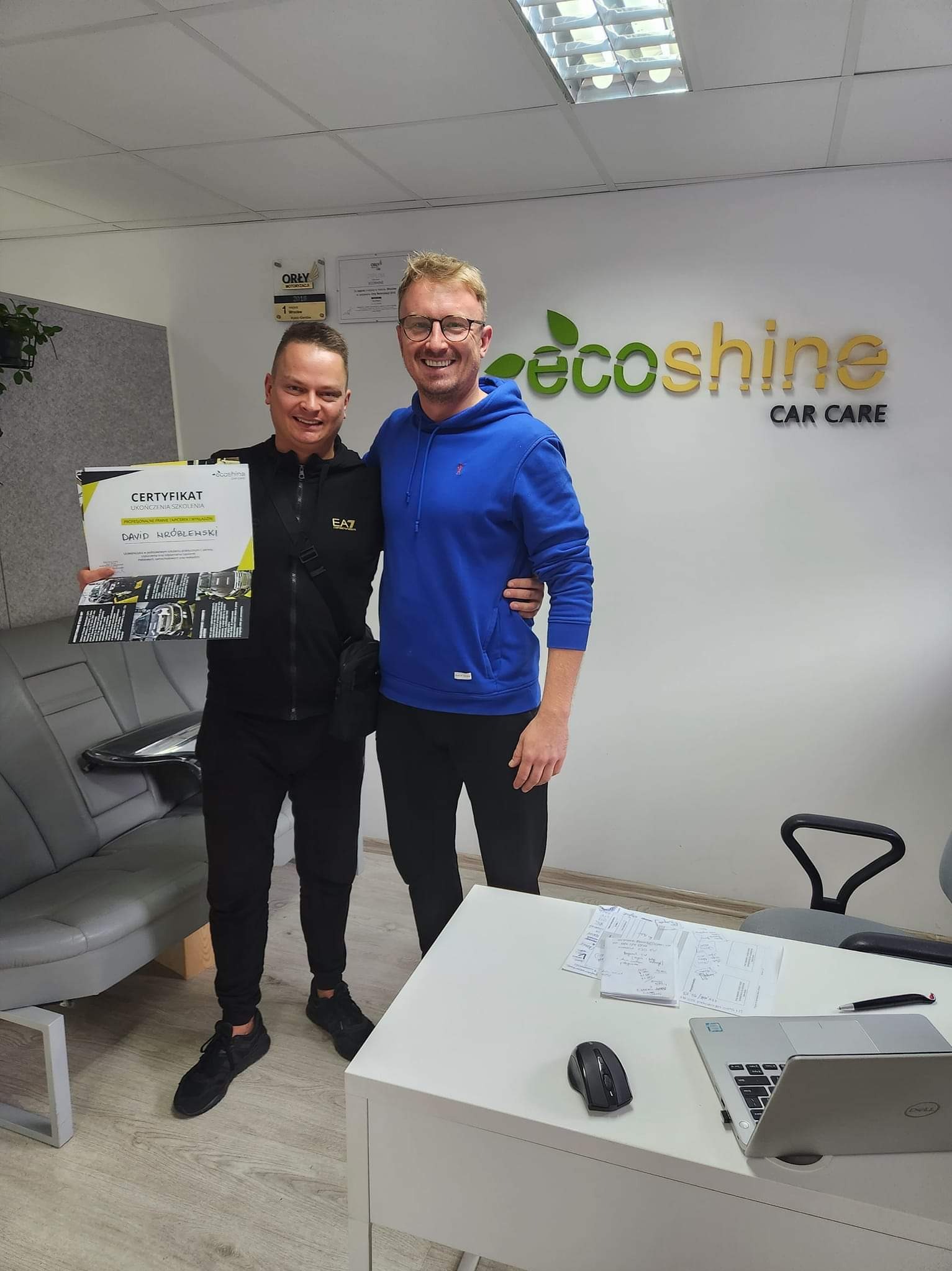 Dwaj mężczyźni pozują z certyfikatem ukończenia szkolenia z profesjonalnego czyszczenia tapicerki samochodowej, na tle biura firmy EcoShine Car Care.