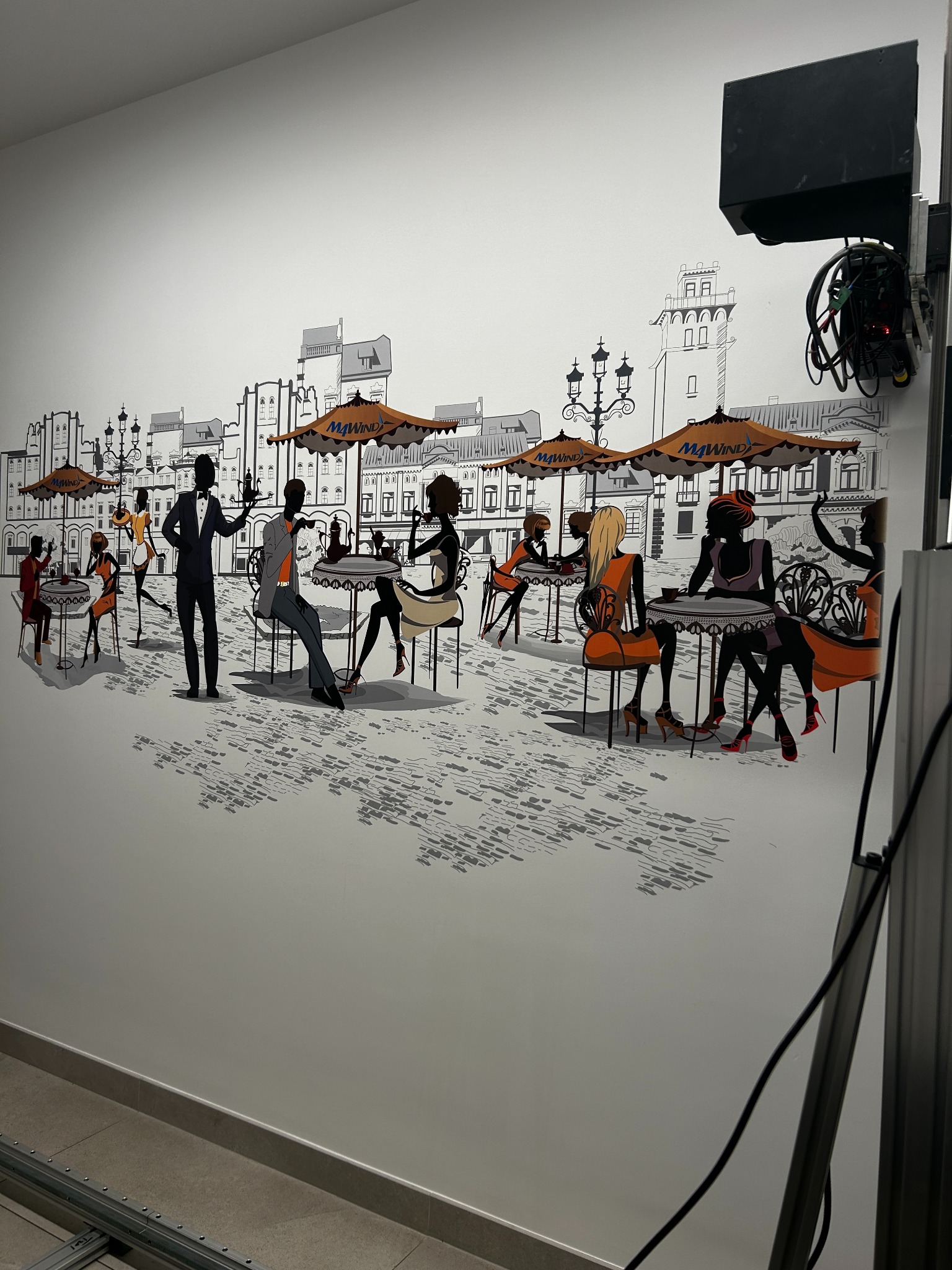 Ilustracja na ścianie przedstawiająca scenę kawiarnianą z sylwetkami ludzi siedzących przy stolikach pod parasolami na tle zarysów budynków miejskich.