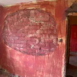 Fragment ściany z odsłoniętą czerwoną cegłą w kształcie owalu, otoczony świeżo nałożoną czerwoną farbą, widoczne zacieki i niedomalowania, surowy stan wykończenia wnętrza.