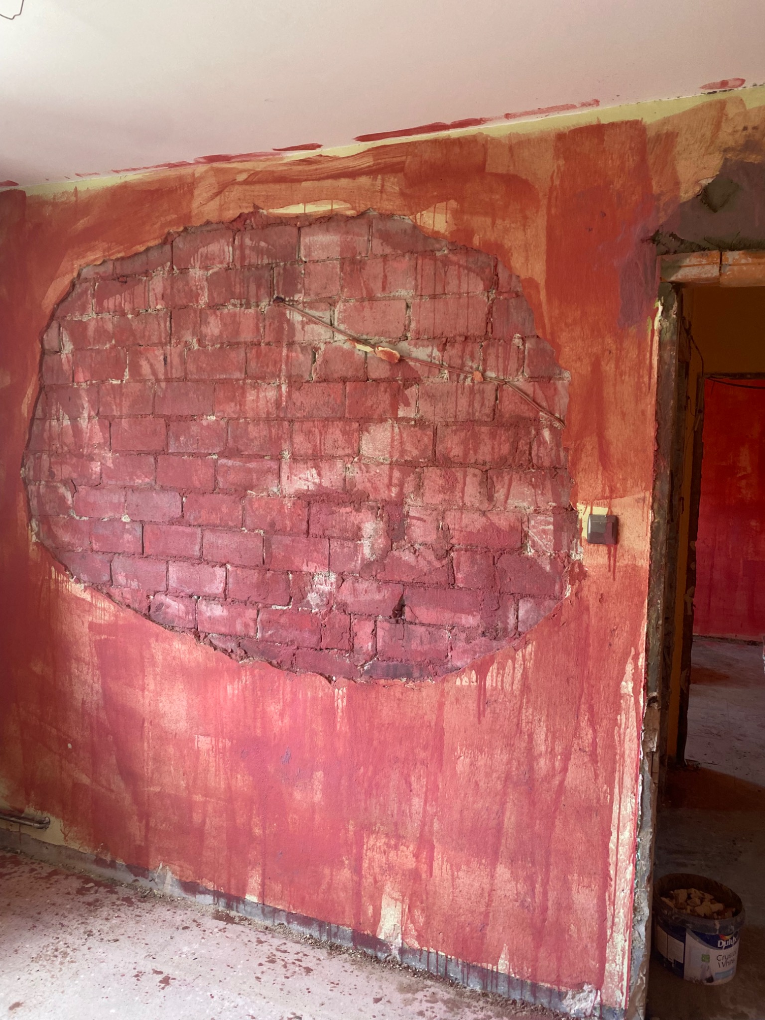 Fragment ściany z odsłoniętą czerwoną cegłą w kształcie owalu, otoczony świeżo nałożoną czerwoną farbą, widoczne zacieki i niedomalowania, surowy stan wykończenia wnętrza.