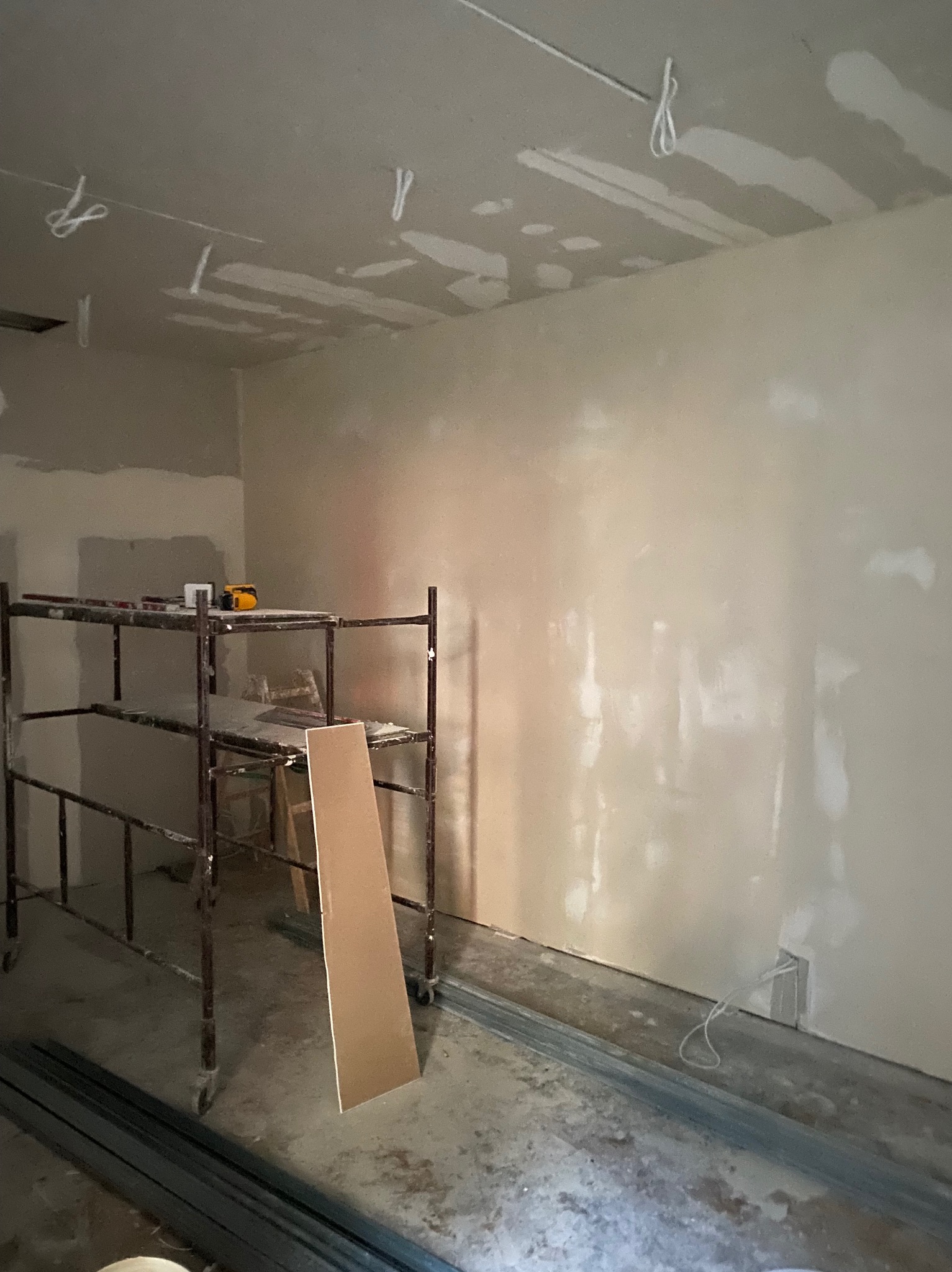 Pokój w trakcie prac wykończeniowych: widoczne szpachlowane ściany, metalowe profile podłogowe, rusztowanie z narzędziami, oraz wystające kable ze ściany i sufitu.