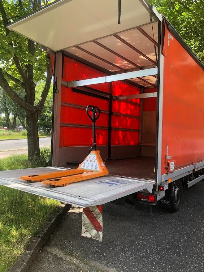 Pomarańczowy wózek paletowy na platformie załadunkowej otwartego busa dostawczego z pomarańczową plandeką, widok z zewnątrz, drzewo obok.