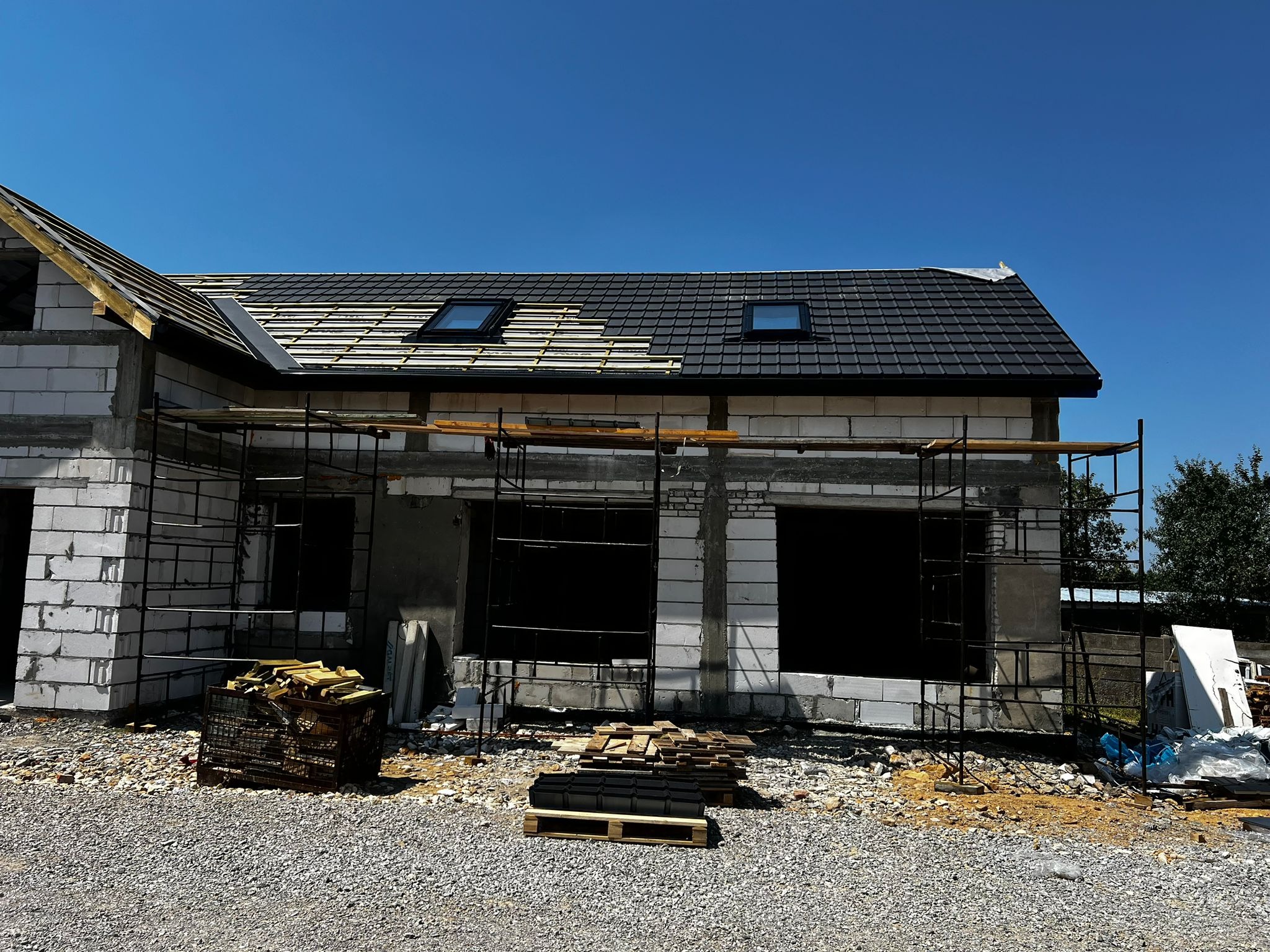 Budowa domu w trakcie realizacji: częściowo pokryty dach z dwoma oknami dachowymi, widoczna więźba dachowa, rusztowania, bloczki betonowe i materiały budowlane na żwirowym podłożu.