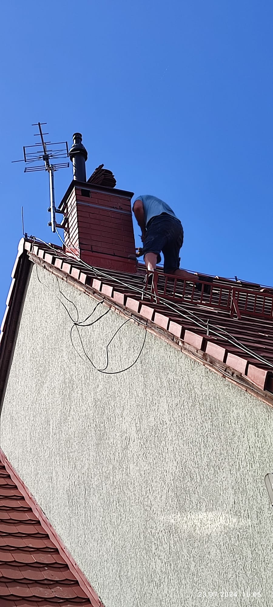 Dekarz pracuje na dachu przy kominie z cegły, widoczne anteny telewizyjne, przewody i drabina dachowa.