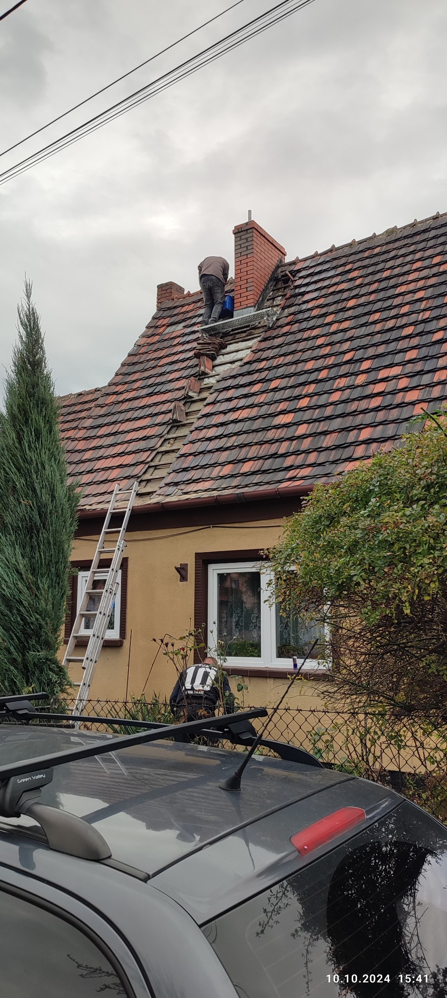 Dekarz na dachu z czerwonej dachówki, częściowo usuniętej w pobliżu komina, zabezpieczony metalową platformą, drabina oparta o ścianę domu, pochmurne niebo.