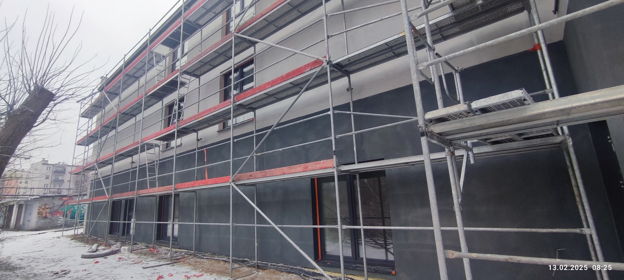 Budynek wielorodzinny w trakcie prac elewacyjnych, osłonięty rusztowaniem; widoczne nowe okna i ocieplenie ścian; w tle drzewo bez liści i fragment zabudowy miejskiej.