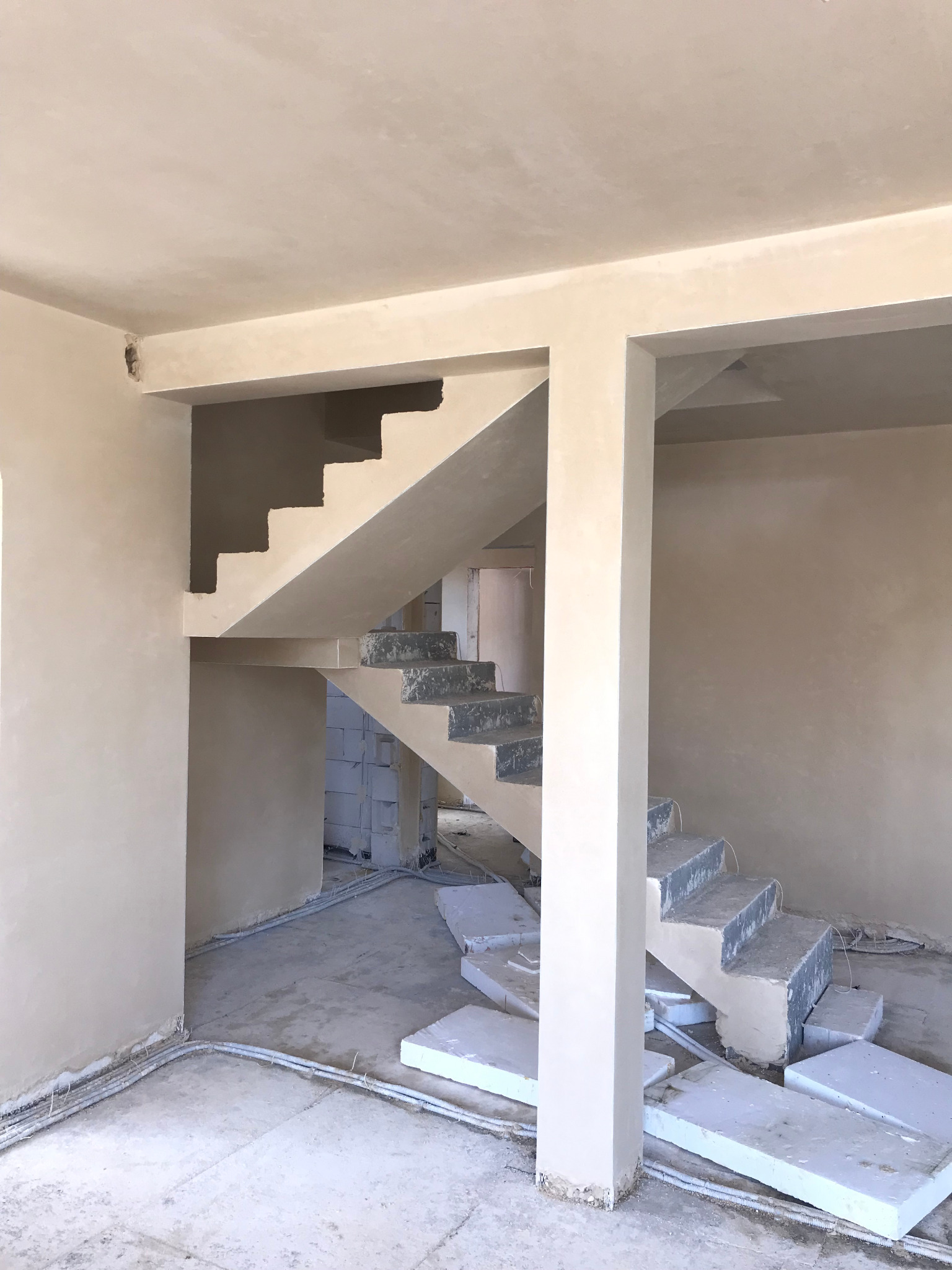 Surowe, jasne wnętrze w trakcie remontu: widoczne surowe tynki na ścianach, betonowe schody w trakcie wykończenia, wystające kable elektryczne oraz porozrzucane płyty styropianowe na podłodze.