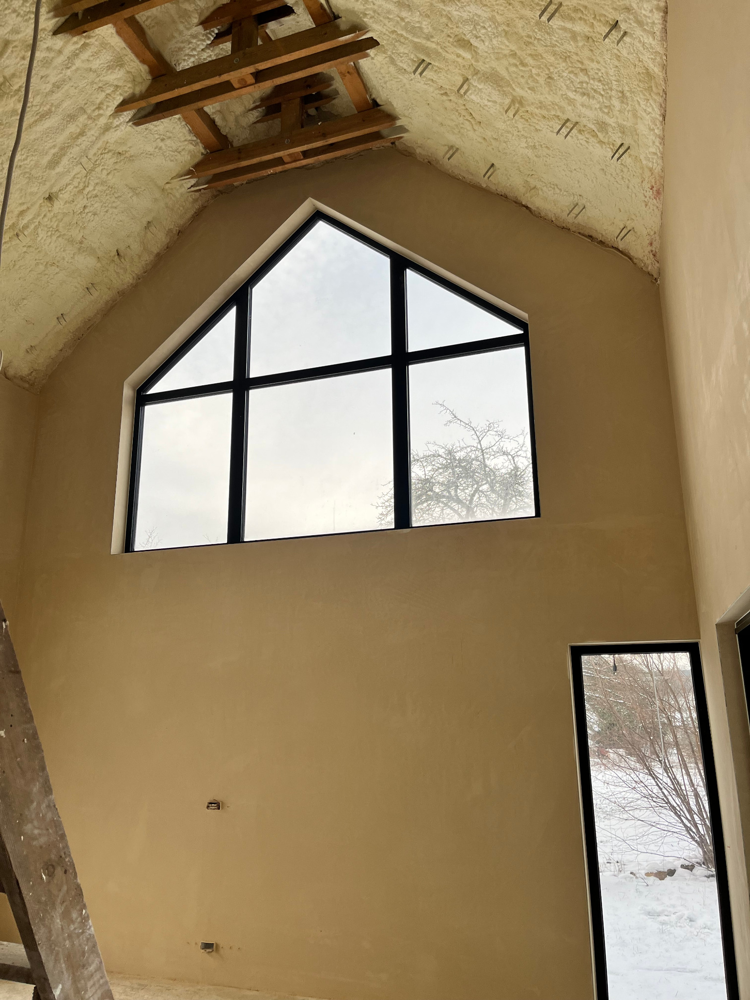 Wnętrze domu w trakcie wykańczania, widoczne ściany pokryte świeżo nałożoną warstwą, duże okno trójkątne z widokiem na zimowy krajobraz, drewniane krokwie i izolacja piankowa na suficie, drabina...