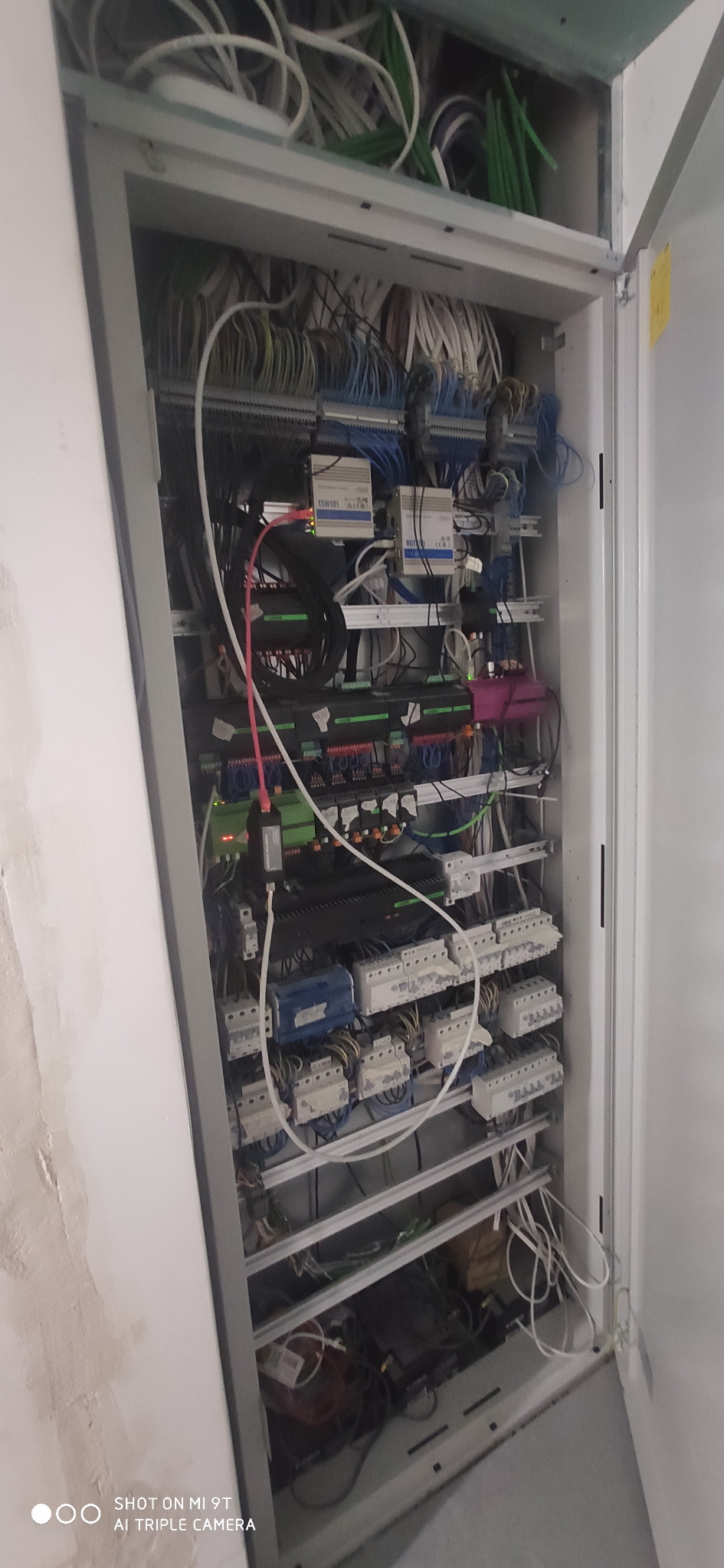 Otwarta szafa sterownicza z widocznymi przewodami, modułami sterującymi, bezpiecznikami i listwami zaciskowymi, w tle widoczna ściana w trakcie wykończenia.