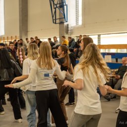 Aleksander Kwiecień - Fotografia - Uczniowie w białych koszulkach tańczą na sali gimnastycznej podczas wydarzenia szkolnego, widoczny baner Ekonomik Szkoła z Pasją oraz logo Liceum Ogólnokształcącego im. Henryka Sienkiewicza...