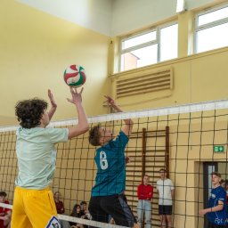Aleksander Kwiecień - Fotografia - Dwóch nastoletnich siatkarzy w trakcie meczu, jeden w koszulce turkusowej z numerem 8 atakuje piłkę nad siatką, drugi w koszulce miętowej próbuje blokować, w tle widownia i żółta ściana sali...