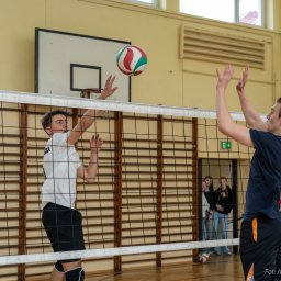 Aleksander Kwiecień - Fotografia - Mecz siatkówki w hali sportowej: dwóch zawodników przy siatce, jeden w białej koszulce atakuje piłkę, drugi w granatowej koszulce próbuje blokować, w tle widoczni kibice i kosz do koszykówki.