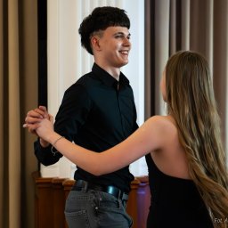 Aleksander Kwiecień - Fotografia - Młoda para tańczy w eleganckim wnętrzu, mężczyzna w czarnej koszuli i jeansach, kobieta w czarnej sukience, oboje uśmiechnięci, widoczne dłonie splecione w tańcu, tło stanowią zasłony i fragment...