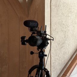 Aleksander Kwiecień - Fotografia - Profesjonalny aparat Sony z mikrofonem Rode na statywie, słuchawki zawieszone na statywie, tło stanowią architektoniczne elementy wnętrza.