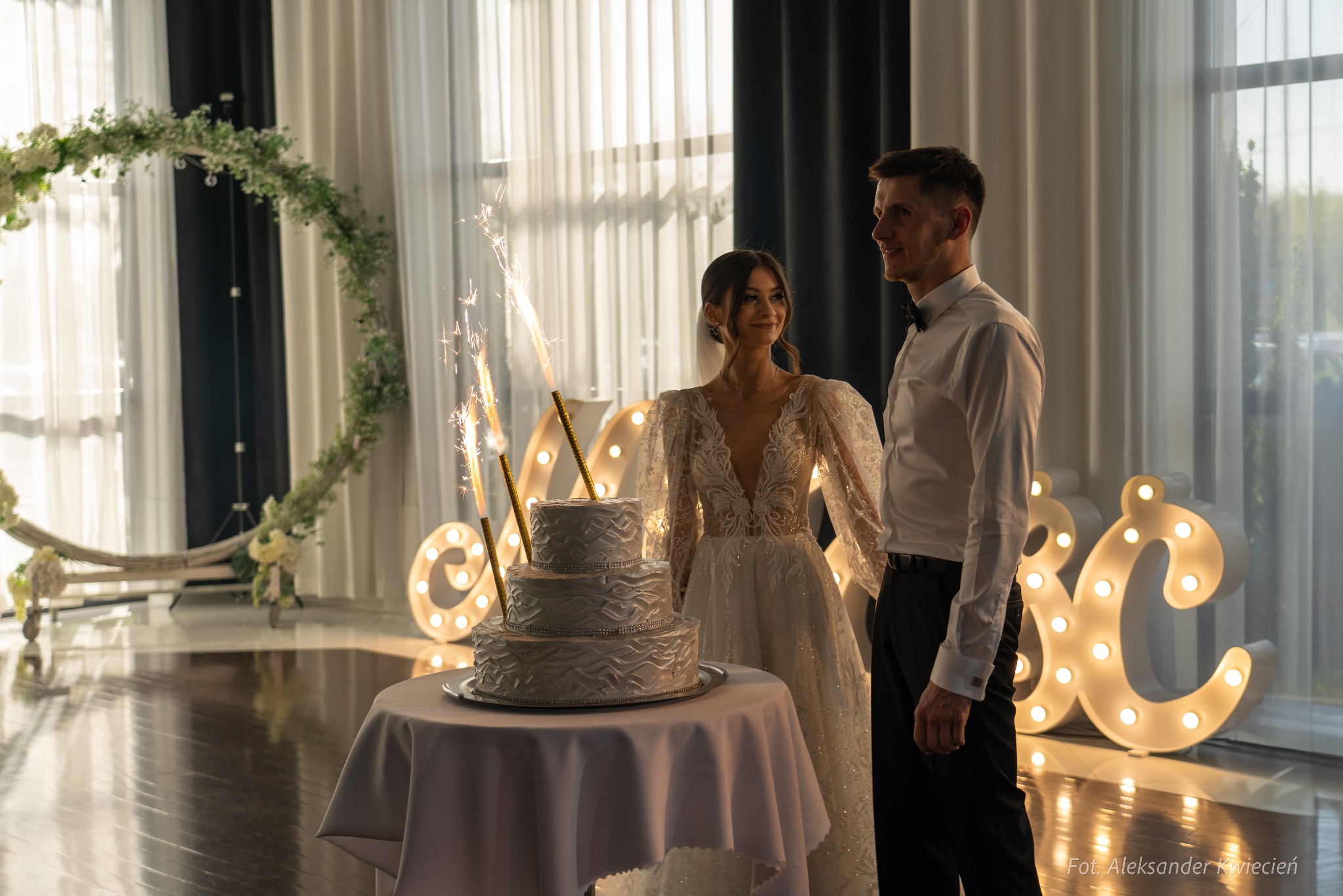 Elegancko ubrana para młoda stoi obok tortu weselnego z fontannami iskier, na tle dekoracji kwiatowych i podświetlanych liter.