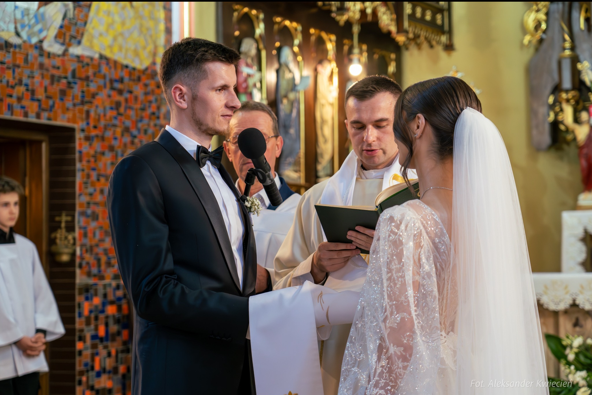 Para młoda podczas ceremonii ślubnej w kościele, ksiądz czyta z księgi, w tle mikrofon i starszy mężczyzna.