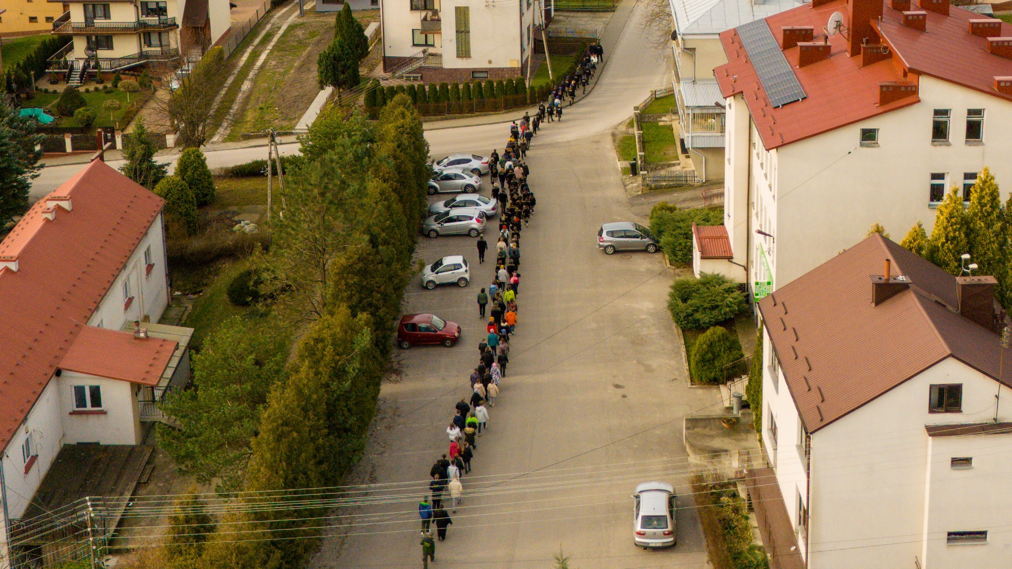 Długi sznur ludzi idzie ulicą między zaparkowanymi samochodami i budynkami, widok z góry, pochmurna pogoda.