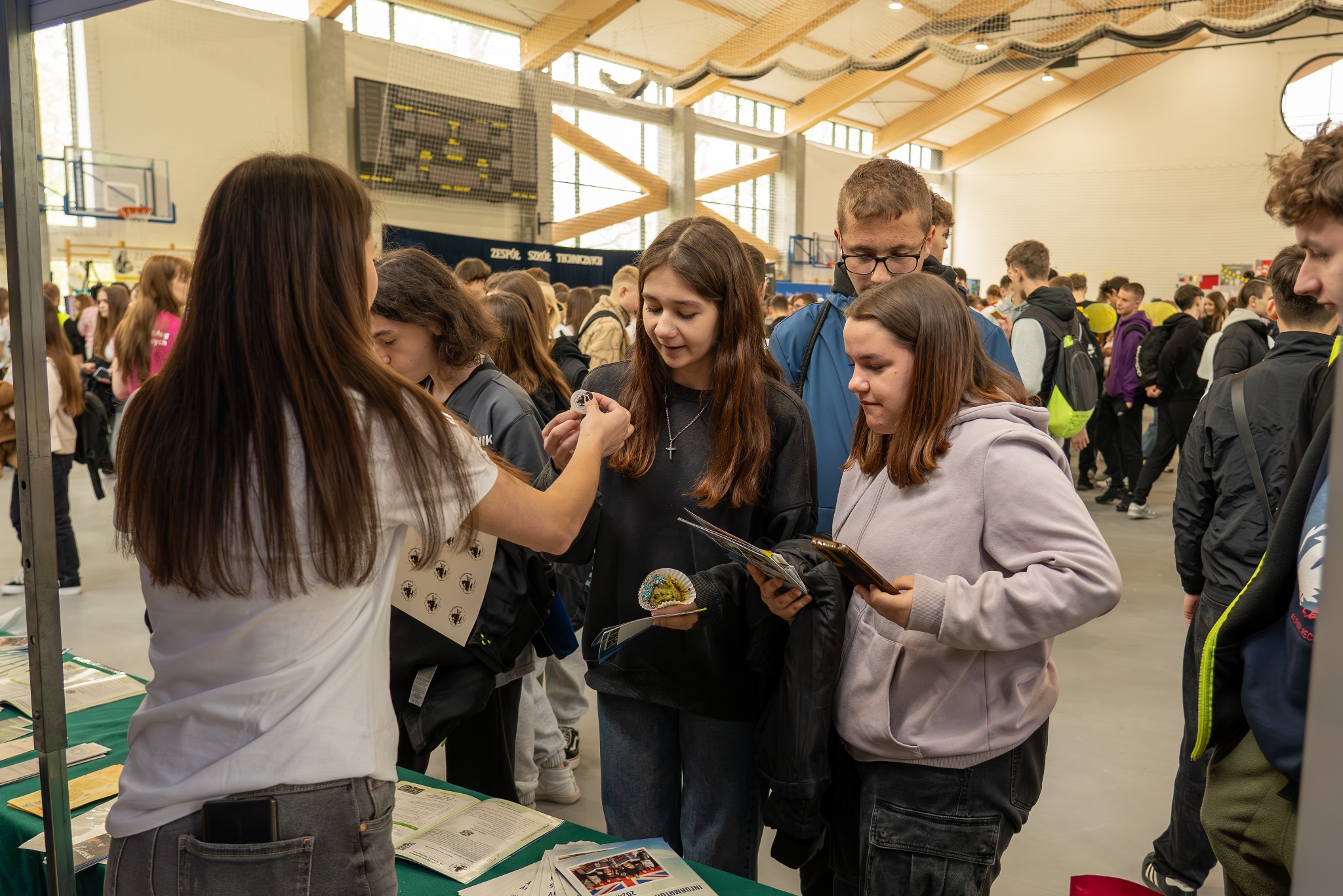 Targi edukacyjne: Uczennica prezentuje medalion z logo szkoły innym uczniom, na stole leżą ulotki informacyjne, w tle hala sportowa z siatką zabezpieczającą.