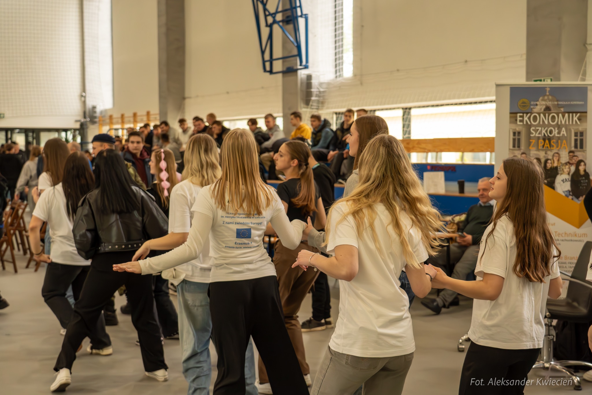 Uczniowie w białych koszulkach tańczą na sali gimnastycznej podczas wydarzenia szkolnego, widoczny baner Ekonomik Szkoła z Pasją oraz logo Liceum Ogólnokształcącego im. Henryka Sienkiewicza...