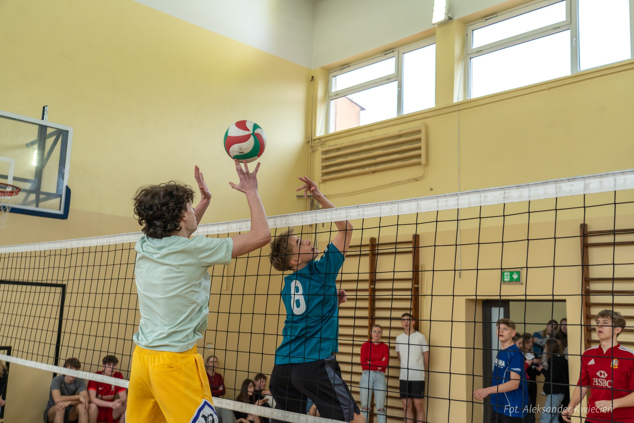 Dwóch nastoletnich siatkarzy w trakcie meczu, jeden w koszulce turkusowej z numerem 8 atakuje piłkę nad siatką, drugi w koszulce miętowej próbuje blokować, w tle widownia i żółta ściana sali...