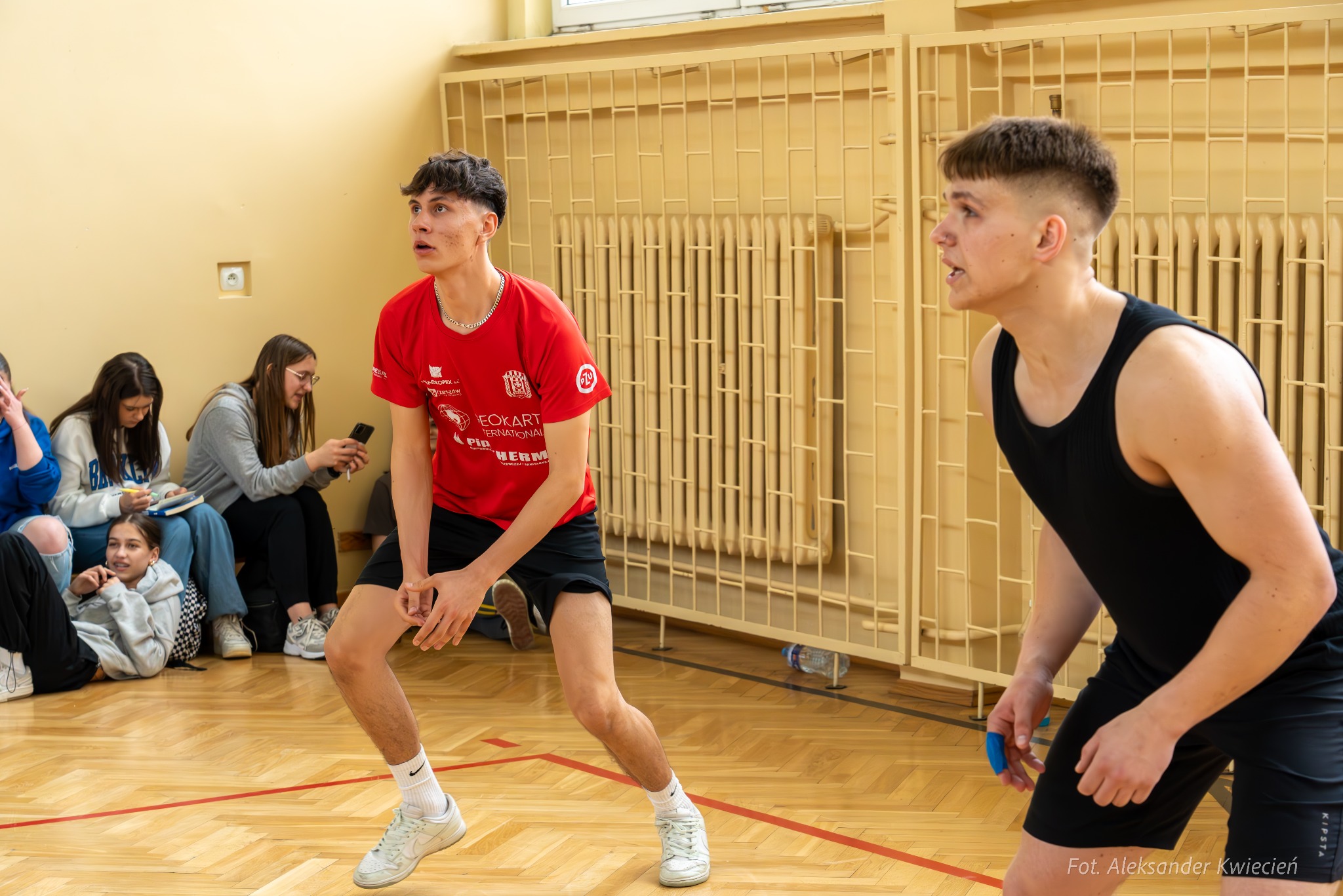 Dwóch młodych mężczyzn gra w siatkówkę na sali gimnastycznej, w tle siedzą obserwatorzy.