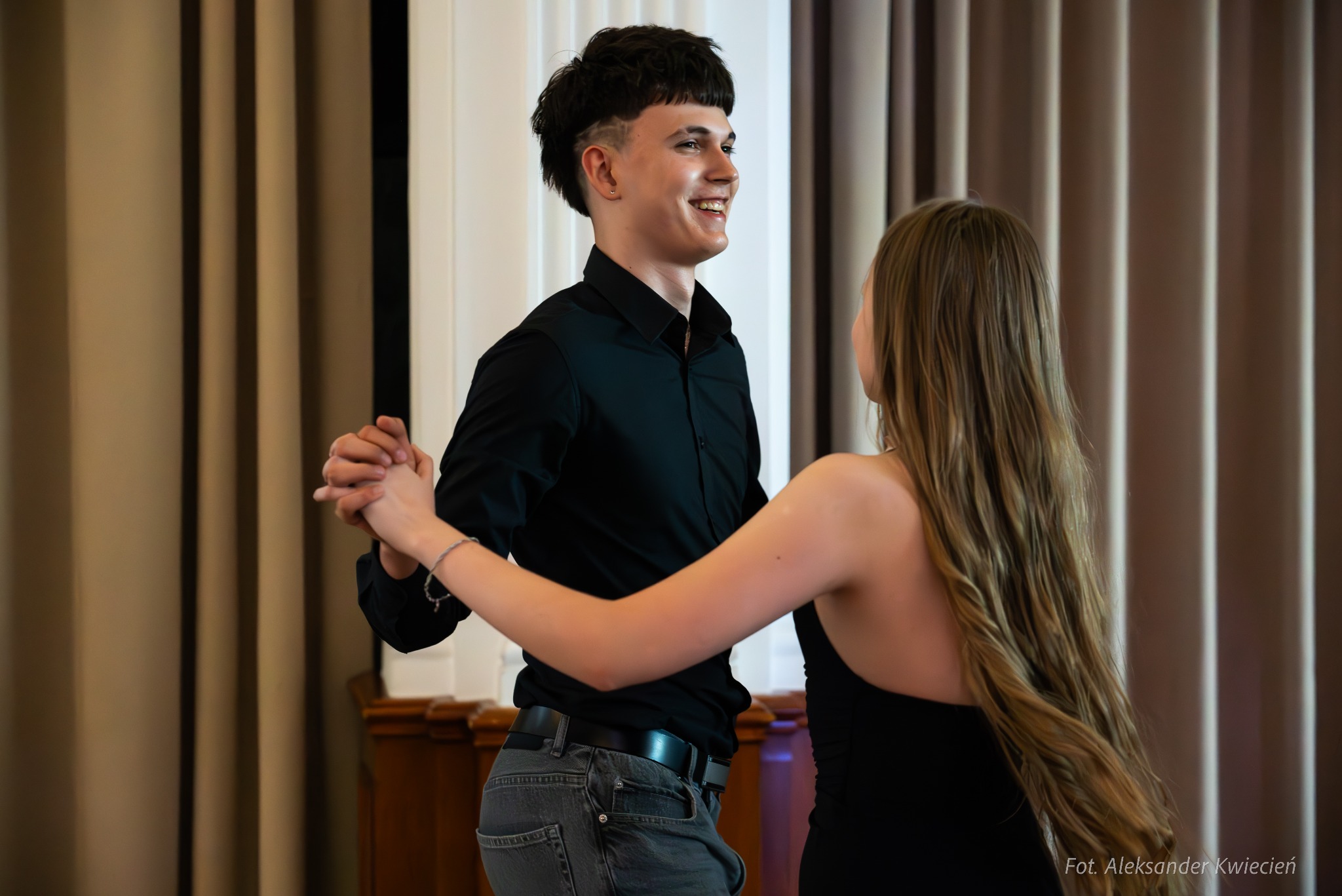 Młoda para tańczy w eleganckim wnętrzu, mężczyzna w czarnej koszuli i jeansach, kobieta w czarnej sukience, oboje uśmiechnięci, widoczne dłonie splecione w tańcu, tło stanowią zasłony i fragment...