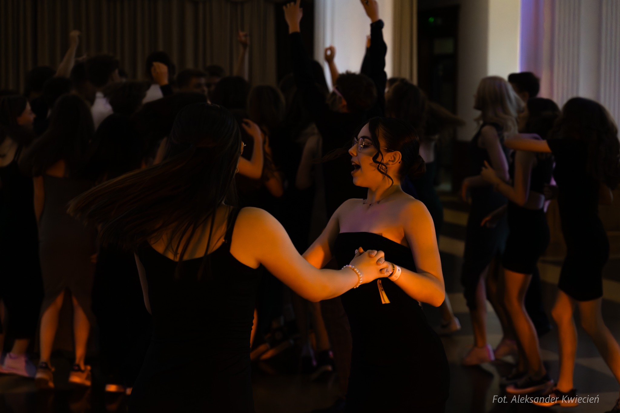 Młode kobiety w czarnych sukienkach tańczące na parkiecie podczas eventu, w tle rozmazane sylwetki innych uczestników.