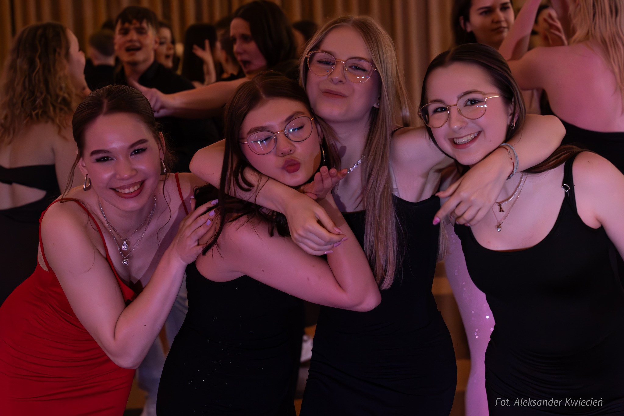 Cztery uśmiechnięte dziewczyny w sukienkach pozują do zdjęcia podczas imprezy, w tle widoczni inni uczestnicy zabawy.