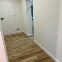 Korytarz z panelami podłogowymi imitującymi drewno, białe ściany z listwami przypodłogowymi, białe drzwi z czarną klamką i widok na dalszą część mieszkania.