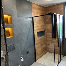 Adrian Pietrucha - Nowoczesna łazienka z kabiną typu walk-in, szare płytki i drewnopodobne panele ścienne. Wnęki z oświetleniem i czarna armatura. Minimalistyczny design.