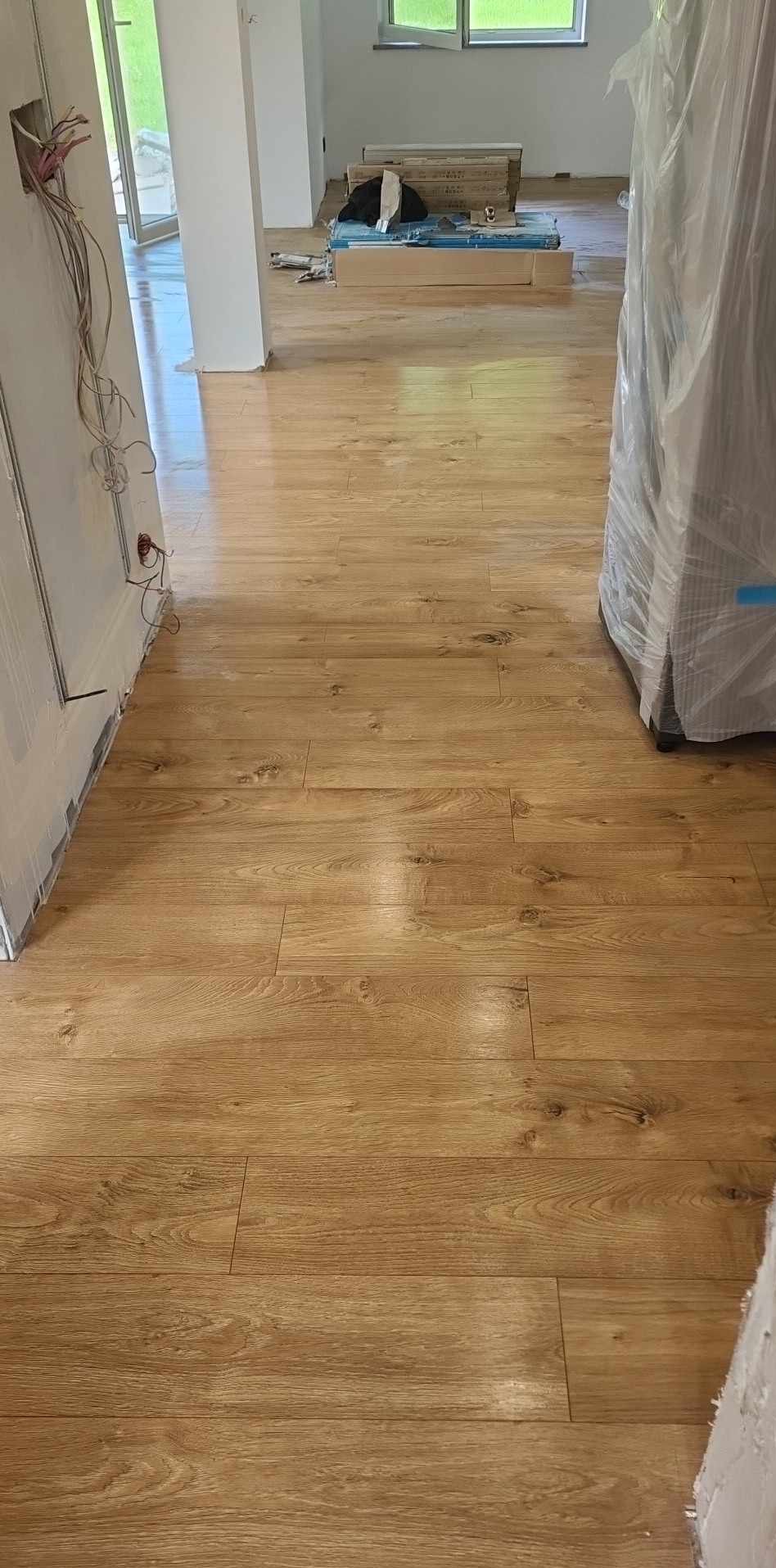 Jasne, drewnopodobne panele podłogowe ułożone w nowym pomieszczeniu, widoczne przewody elektryczne wystające ze ściany i materiały budowlane w tle.